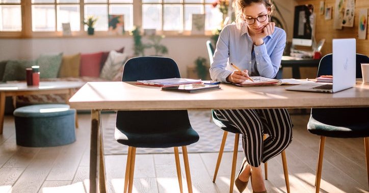 A woman sitting at a desk in a sunny loft and sketching in a notebook next to an open laptop.
