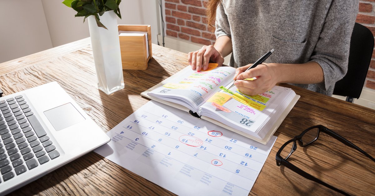 A women sitting at a sunny desk with a laptop, calendar, and planner.