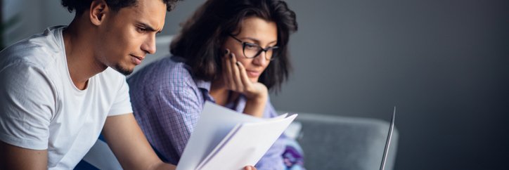 A worried-looking man and woman sitting on their couch and going through paper bills with a laptop open on the coffee table.