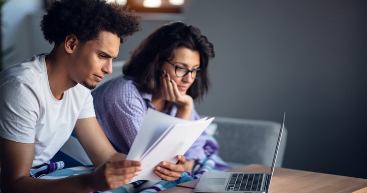 A worried-looking man and woman sitting on their couch and going through paper bills with a laptop open on the coffee table.