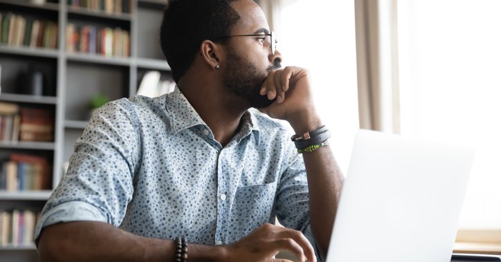 A person sitting at a desk in front of an open laptop and glancing out the window to his left.