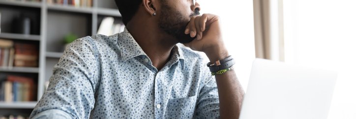 A person sitting at a desk in front of an open laptop and glancing out the window to his left.