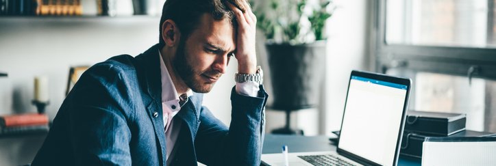 A man sitting at his office desk and looking stressed with his hand on his forehead while he fills in paperwork.
