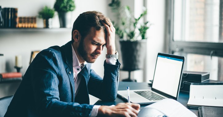 A man sitting at his office desk and looking stressed with his hand on his forehead while he fills in paperwork.