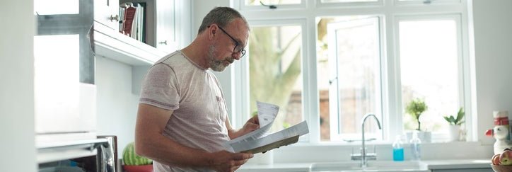 A worried-looking man standing in his kitchen and reading through paper bills in his hands.