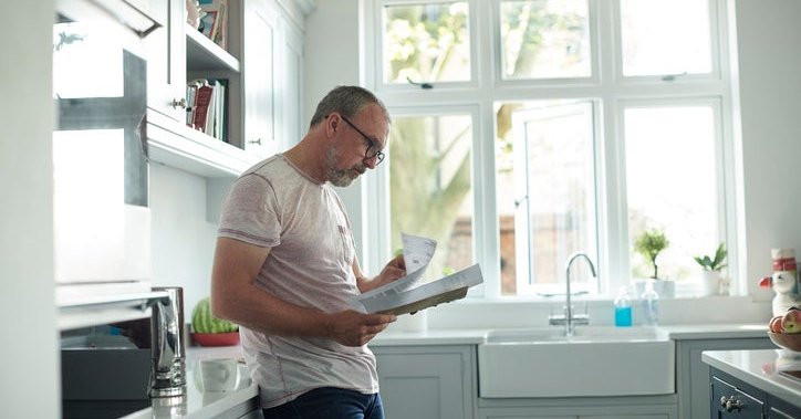 A worried-looking man standing in his kitchen and reading through paper bills in his hands.