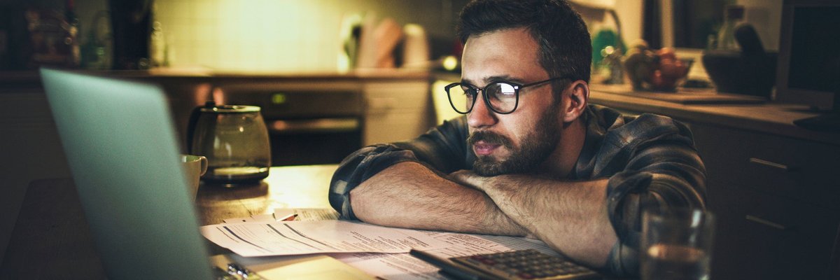 A person looking worriedly at a laptop with their head resting on their arms on the kitchen counter.