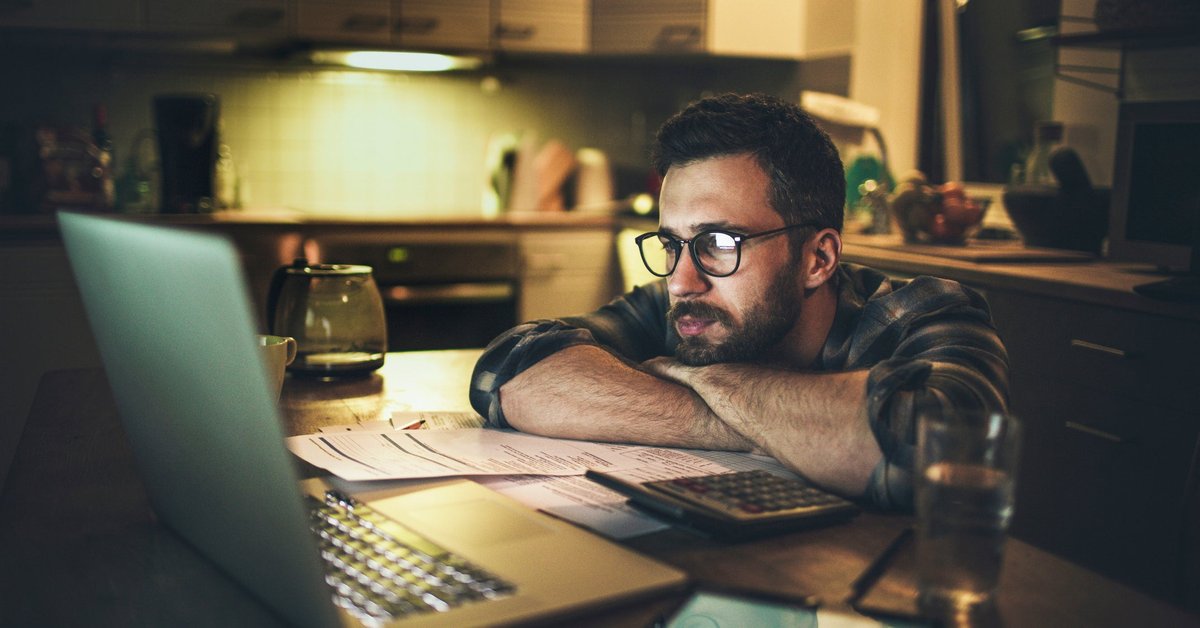A person looking worriedly at a laptop with their head resting on their arms on the kitchen counter.