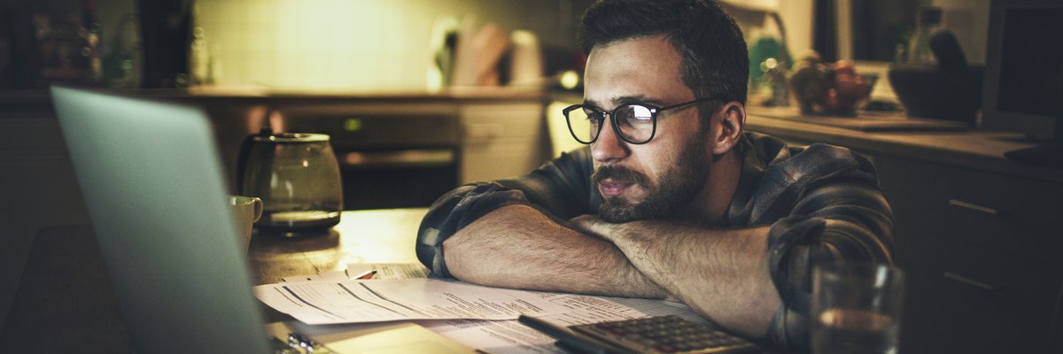A person looking worriedly at a laptop with their head resting on their arms on the kitchen counter.