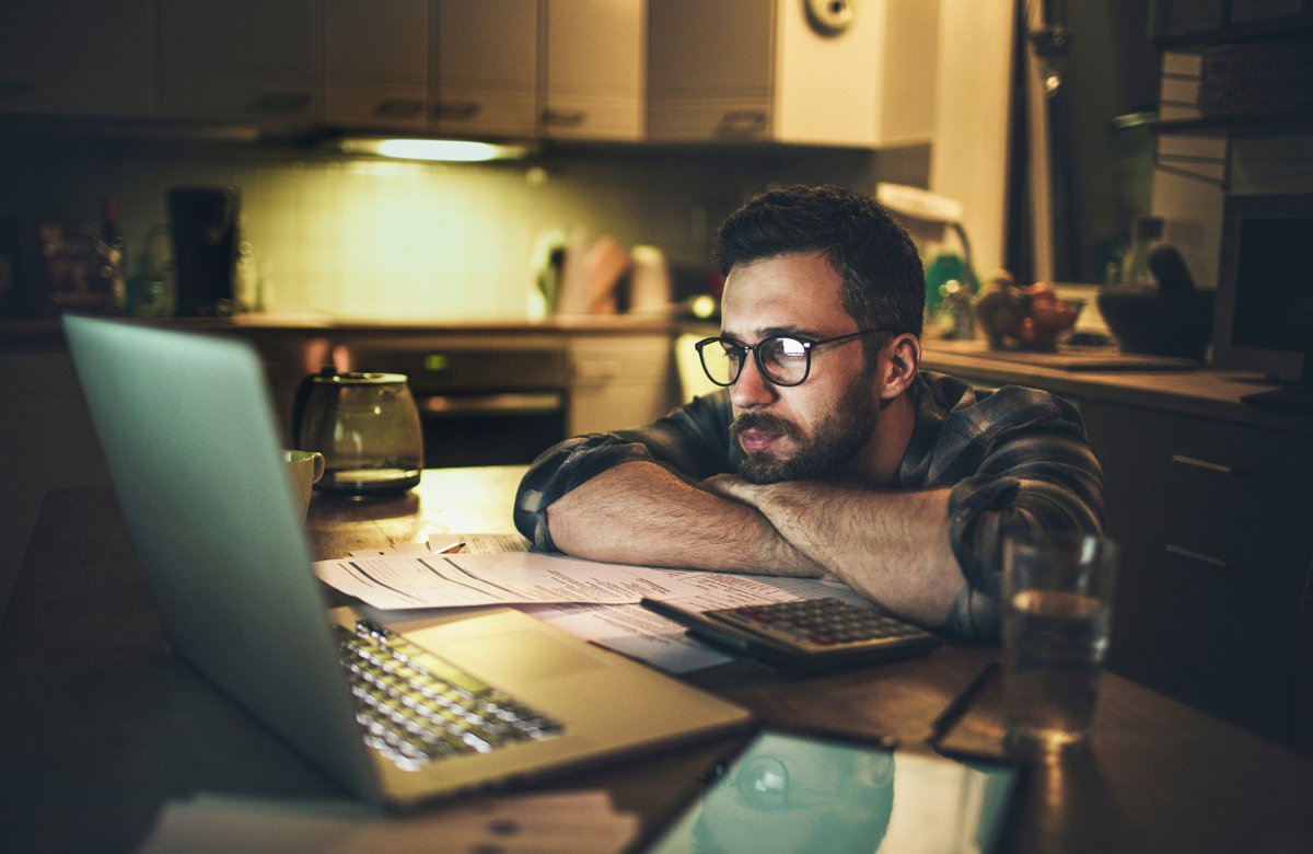 A person looking anxiously at a laptop with his head resting in his arms on the kitchen table.