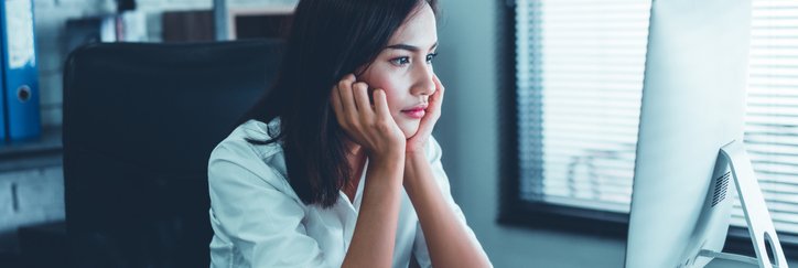 A woman sitting at a desk in front of a computer and staring into space with her chin resting in her hands.