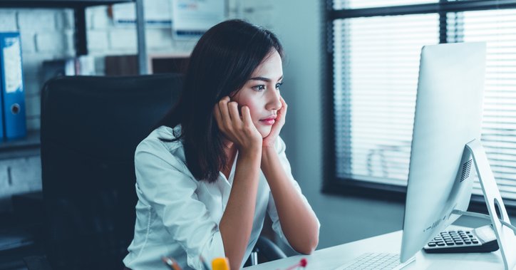 A woman sitting at a desk in front of a computer and staring into space with her chin resting in her hands.