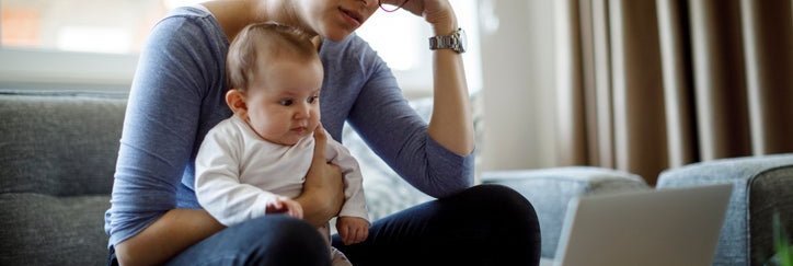 A worried woman sitting on her couch with her baby on her lap while she looks at her laptop.