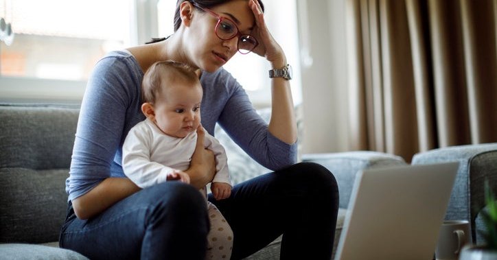 A worried woman sitting on her couch with her baby on her lap while she looks at her laptop.