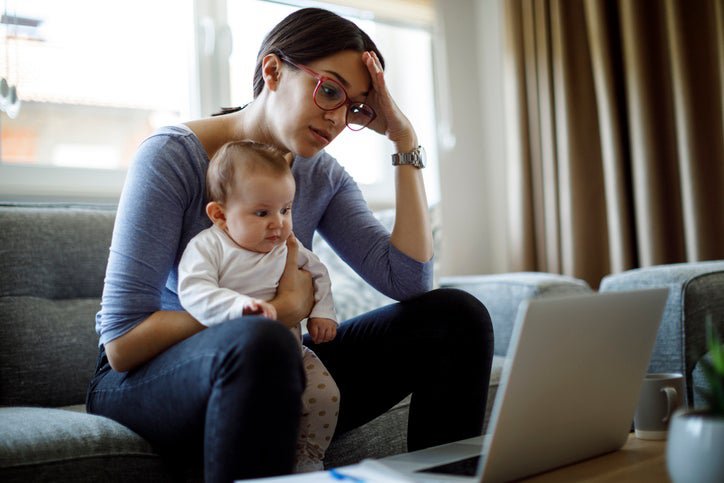 A worried woman sitting on her couch with her baby on her lap while she looks at her laptop.