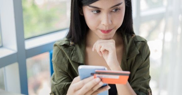 young woman holding credit card and smartphone and thinking with chin on fist