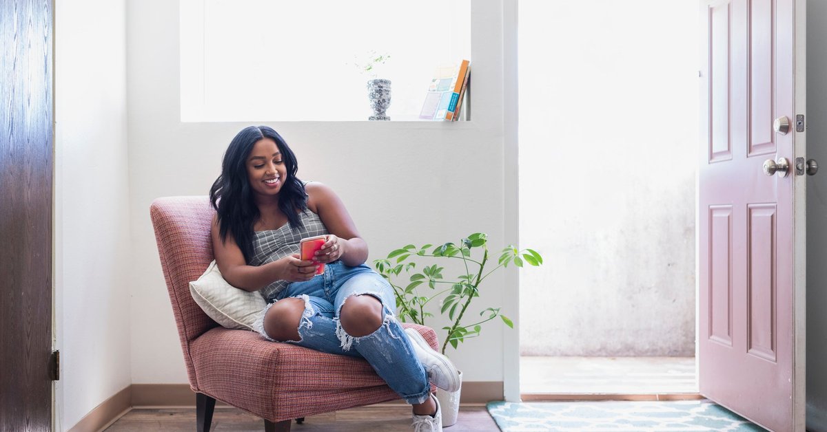 A smiling young adult sitting in an armchair in a sunny room while looking at a phone.