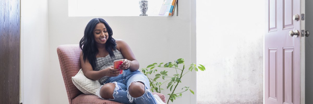 A smiling young adult sitting in an armchair in a sunny room while looking at a phone.