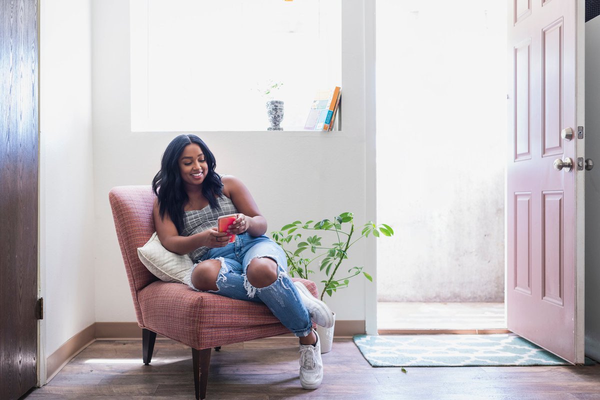 A smiling young adult sitting in an armchair in a sunny room while looking at a phone.