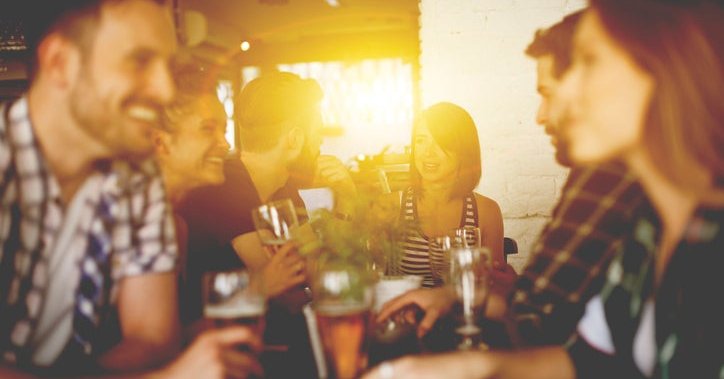 A group of young adults sitting at a table in a sunny bar enjoying beers and chatting.