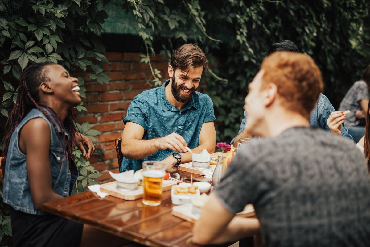 Group of young adults laughing and enjoying drinks and food at a patio table in a restaurant.