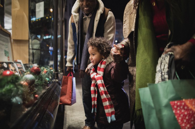 A young child holding hands with his parents and looking at a holiday window display.