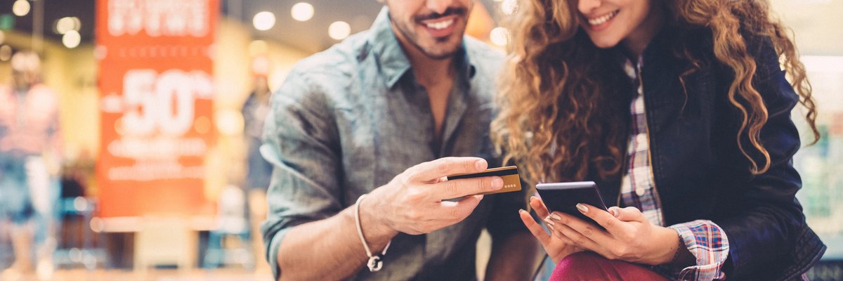 A young couple sitting on a bench at a mall with a phone and credit card in their hands.