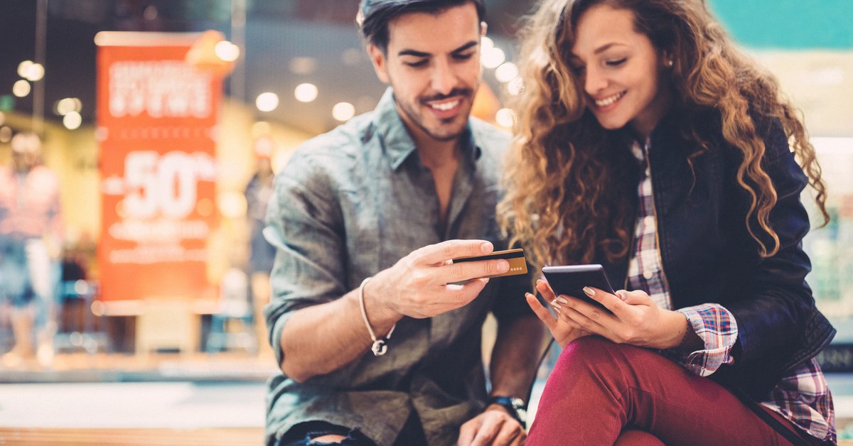 A young couple sitting on a bench at a mall with a phone and credit card in their hands.
