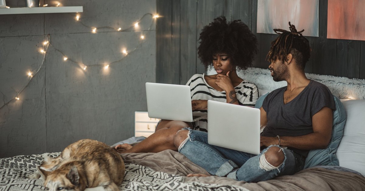 A young couple sitting on their bed with their dog and each shopping online with their laptops.
