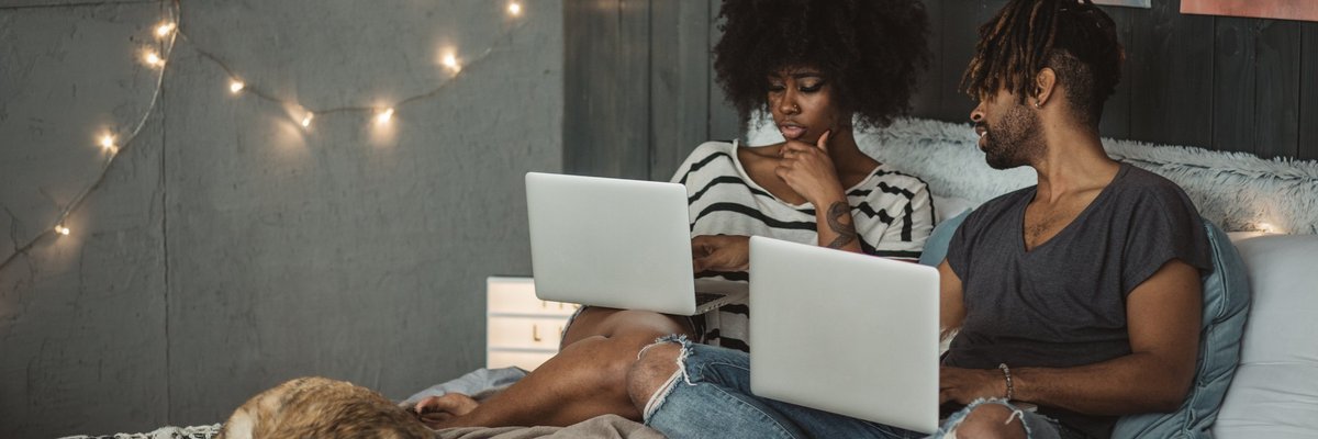 A young couple sitting on their bed with their dog and each shopping online with their laptops.