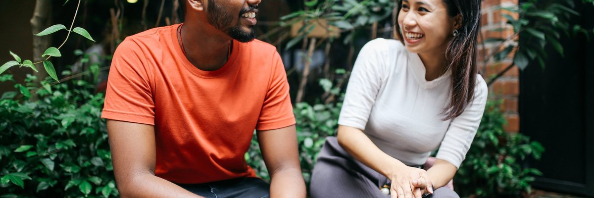 A young couple smiling and chatting while sitting on a brick ledge in front of green bushes.