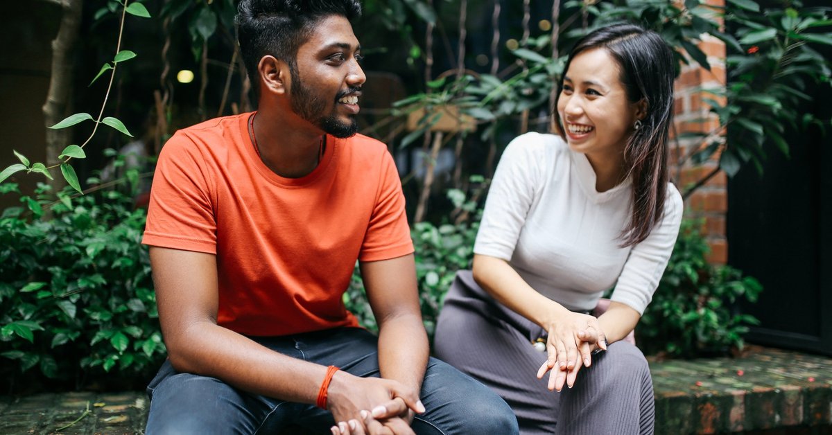 A young couple smiling and chatting while sitting on a brick ledge in front of green bushes.