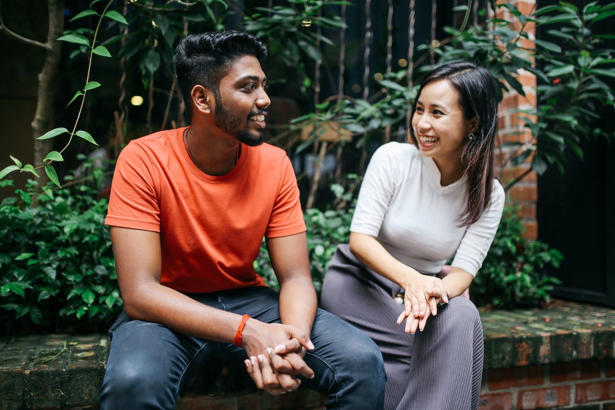 A young couple smiling and chatting while sitting on a brick ledge in front of green bushes.