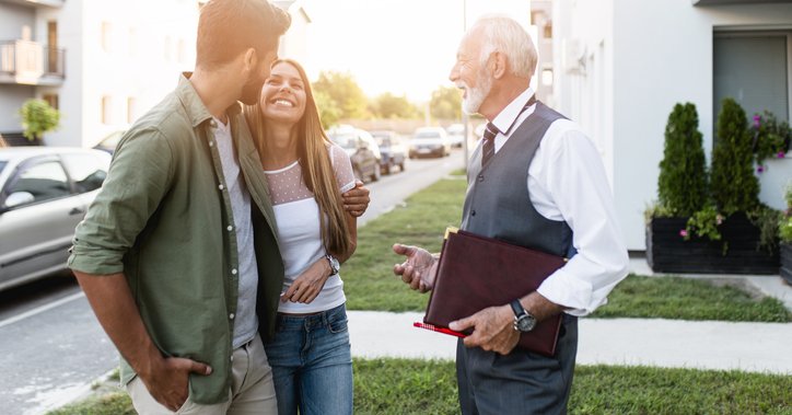 A young couple smiling while talking to their realtor outside a home for sale.