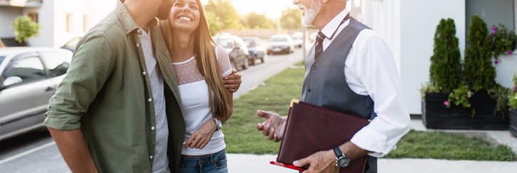 A young couple smiling while talking to their realtor outside a home for sale.