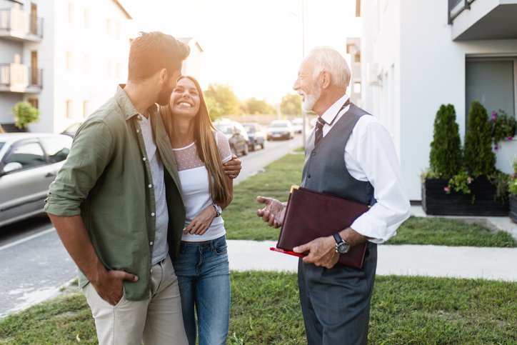 A young couple smiling while talking to their realtor outside a home for sale.