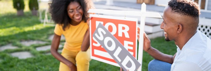 A smiling young couple placing a Sold sign on the For Sale sign in front of their new home.