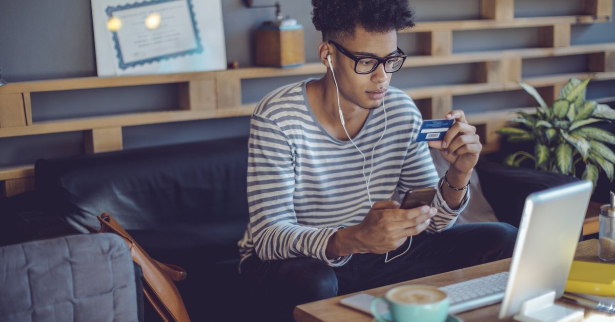 A young man on his couch holding a credit card and his phone.