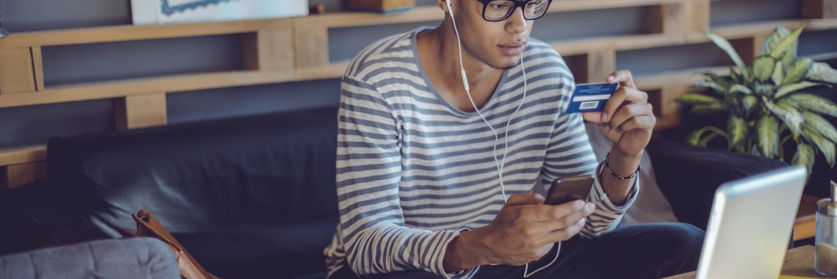A young man on his couch holding a credit card and his phone.