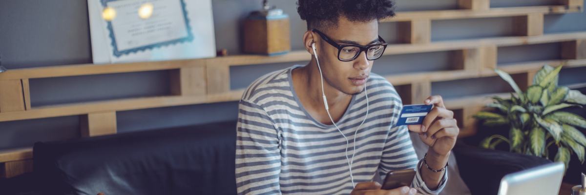 A young man on his couch holding a credit card and his phone.