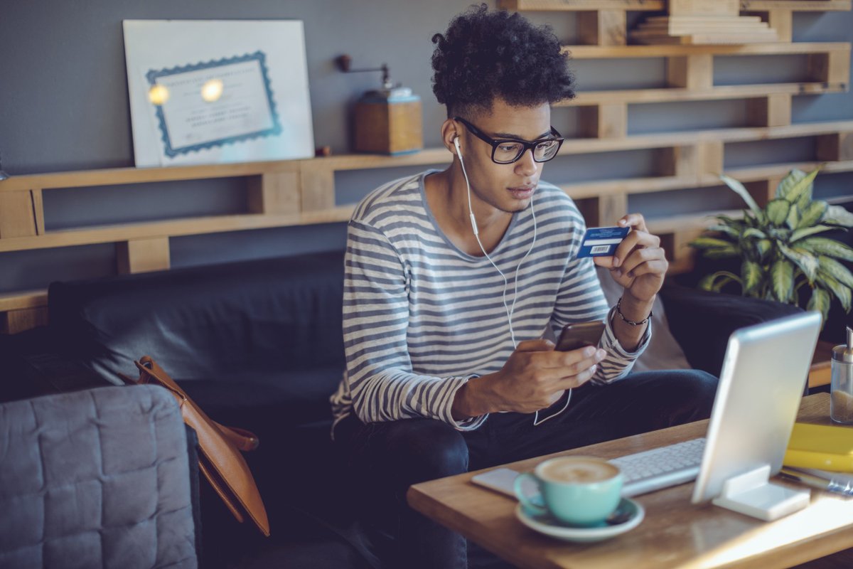 A young man on his couch holding a credit card and his phone.