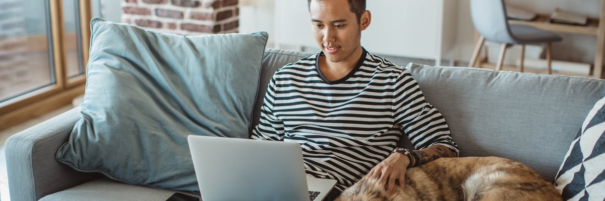 A young man doing work on a laptop while sitting on his couch next to his dog.