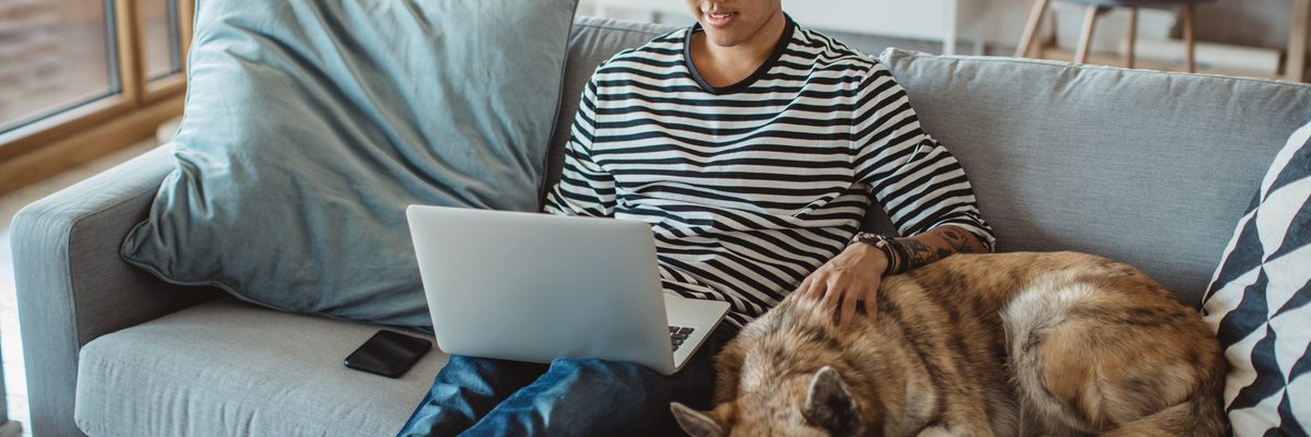 A young man doing work on a laptop while sitting on his couch next to his dog.