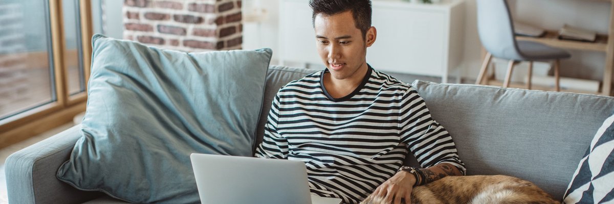 A young man doing work on a laptop while sitting on his couch next to his dog.