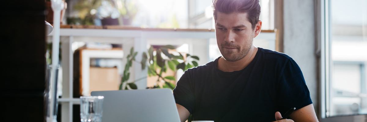 A man sitting at a sunny desk and looking up something on a laptop.