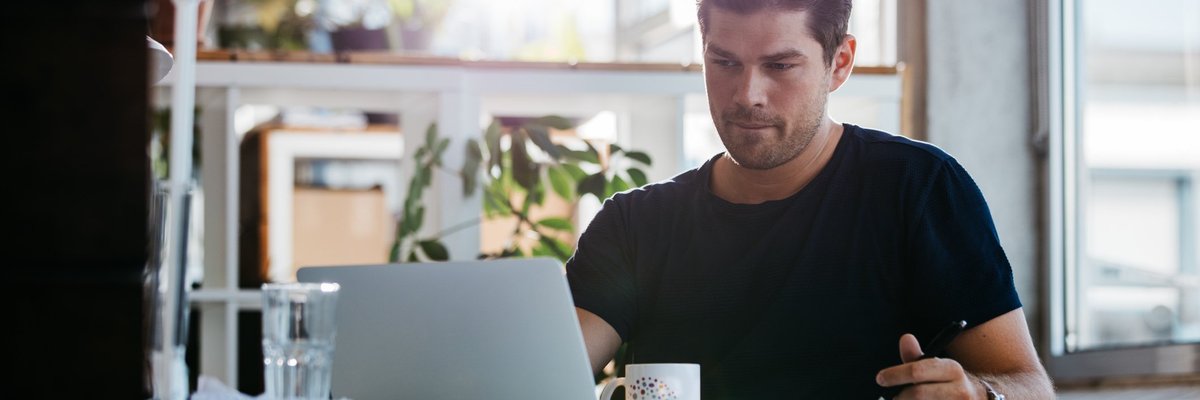 A man sitting at a sunny desk and looking up something on a laptop.
