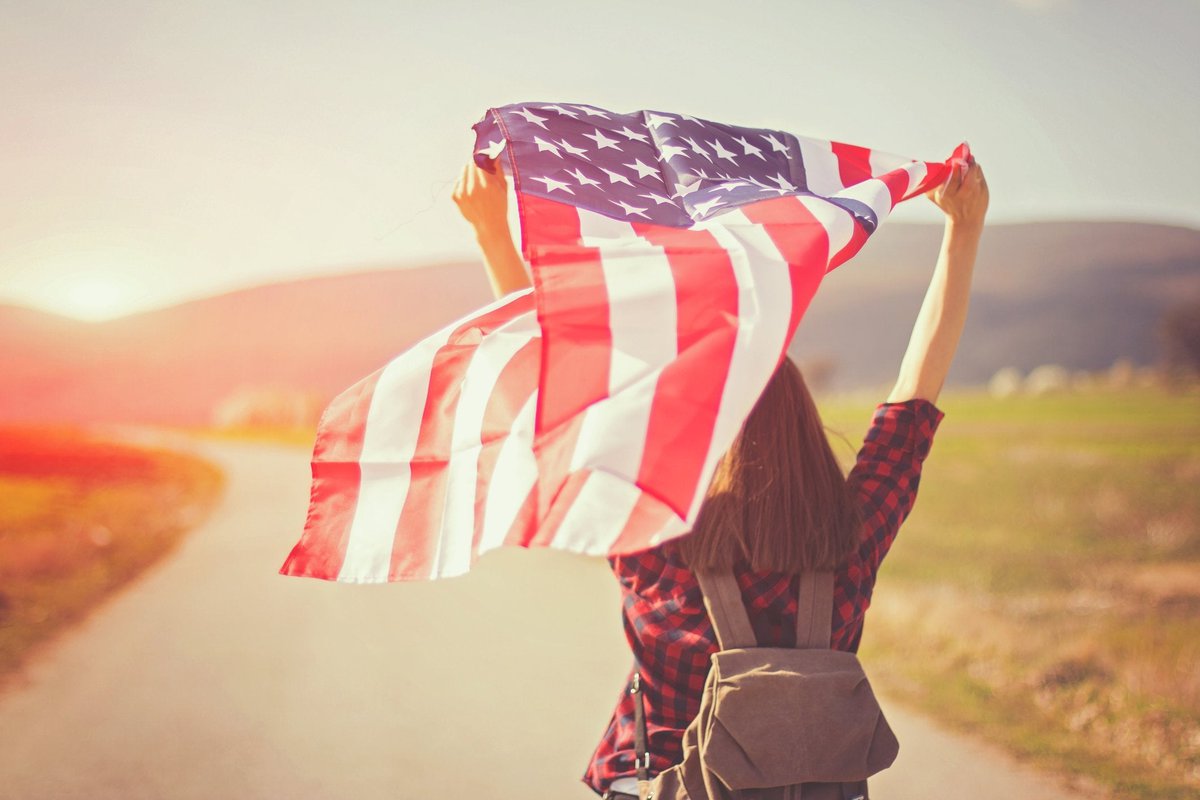 Young woman walking down road with backpack holding American flag.