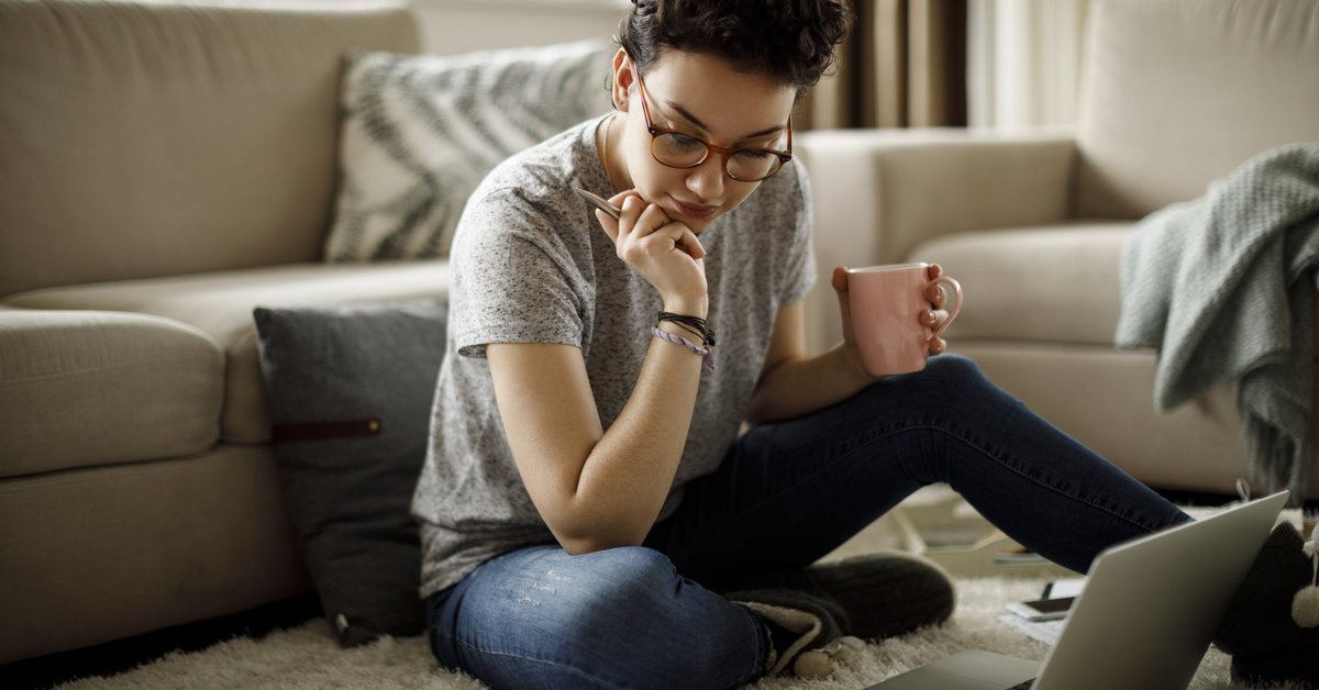 A young woman sitting on the floor of her living room with her laptop and bills around her.