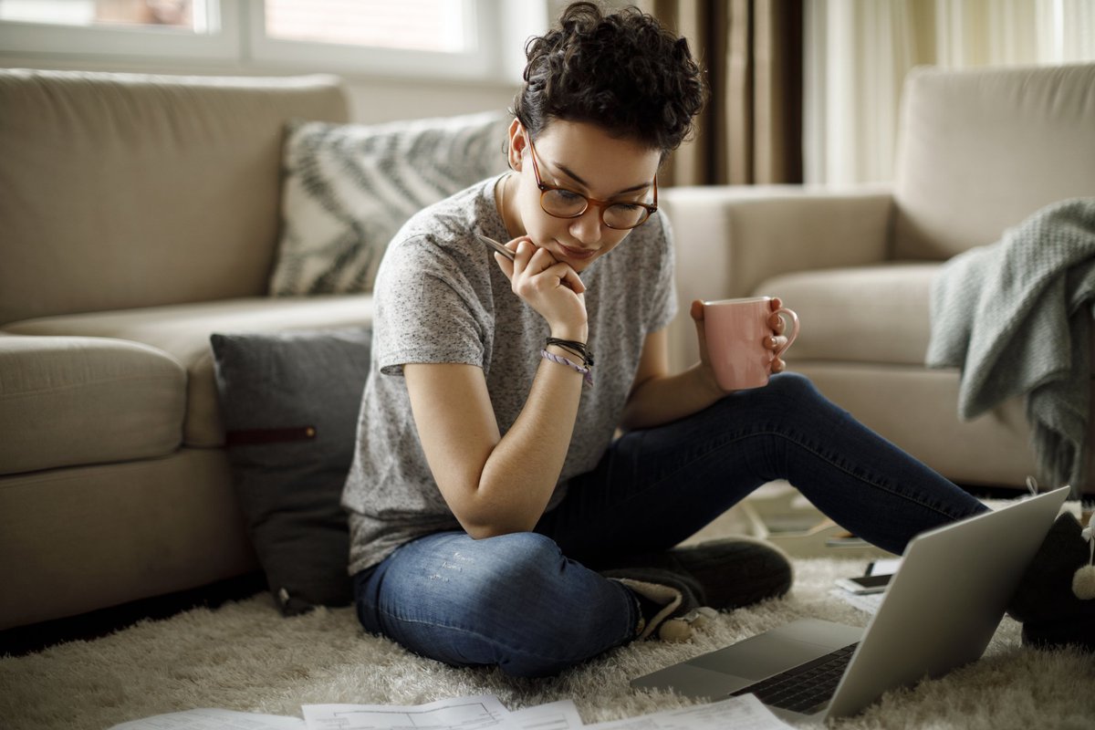 A young woman sitting on the floor of her living room with her laptop and bills around her.