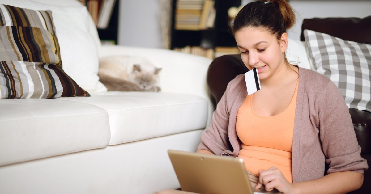 A young woman shops on her laptop holding her credit card while her cat lounges in the background